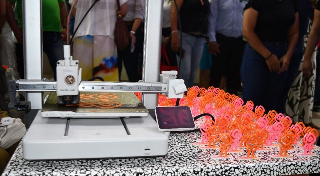 Impresora 3D creando figuras de jóvenes en color naranja y rosa en un evento cultural en Huila, Colombia. Tecnología innovadora y arte se combinan en esta exhibición.