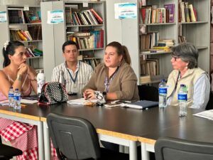 1. Mujeres en reunión en biblioteca, discutiendo temas académicos y culturales, rodeadas de libros en estantes, en un ambiente de aprendizaje y colaboración.