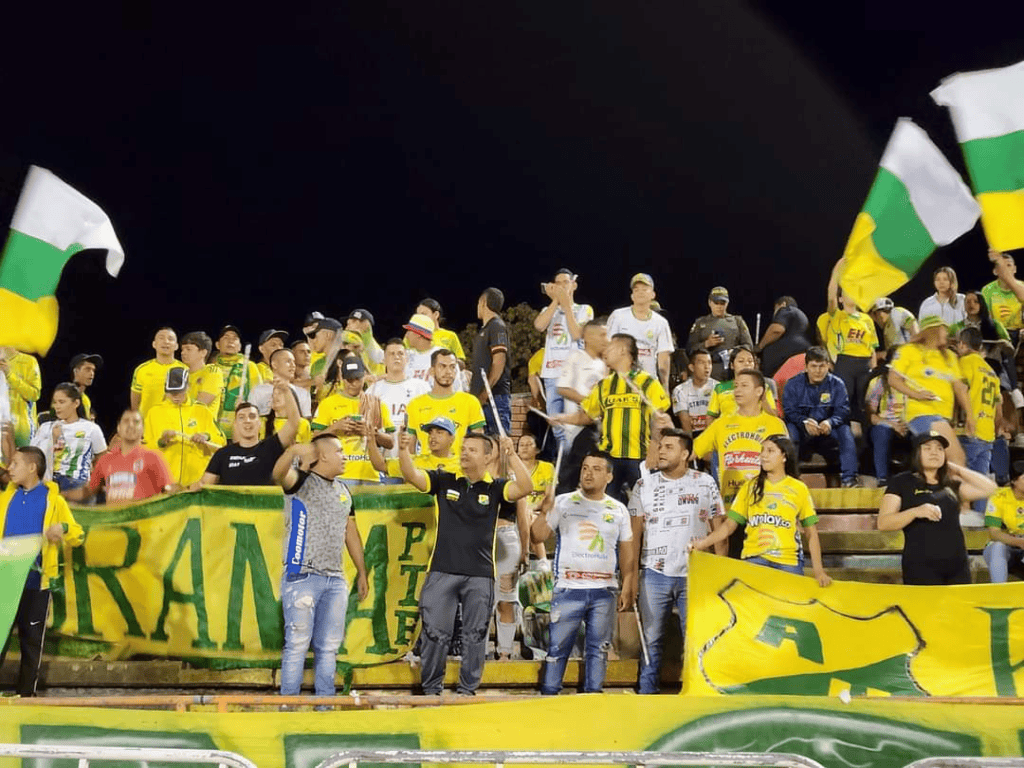 Aficionados del Huila FC en el estadio, mostrando entusiasmo con banderas y camisetas amarillas en apoyo a su equipo durante un partido de fútbol.