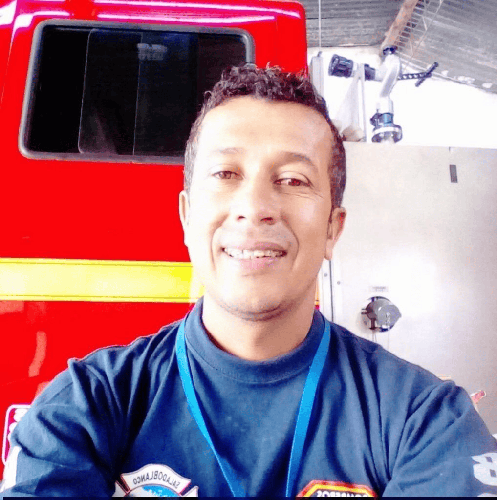 1. Hombre sonriendo en evento de emergencia con unidad de rescate roja y equipo técnico en fondo industrial, fotografía para Huila TV.