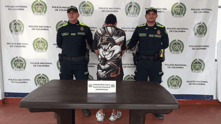 Policía de Colombia detiene a un sospechoso en una rueda de prensa en Huila, con fondo de logotipos oficiales y una mesa en primer plano, en un operativo de seguridad.