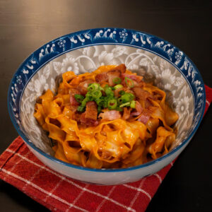 Creamy gochujang noodles topped with crispy bacon and green onions served in a patterned bowl.