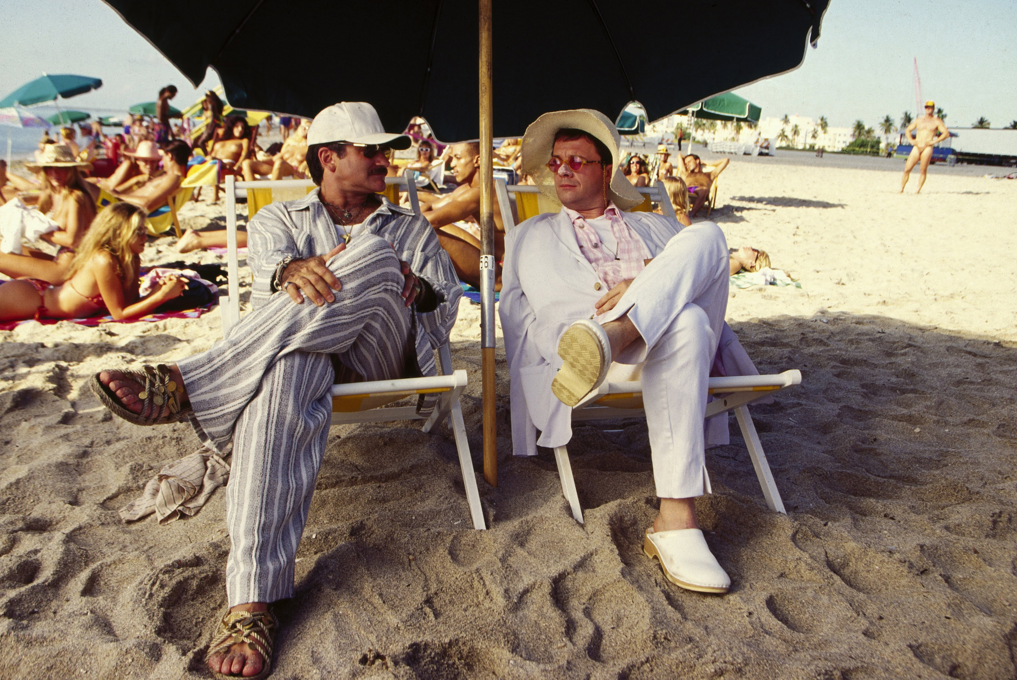Two men sitting under a beach umbrella on a crowded sandy beach.