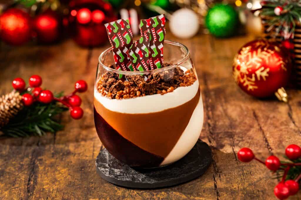 Chocolate layered dessert with festive holiday toppings in a glass, Christmas decorations in background.