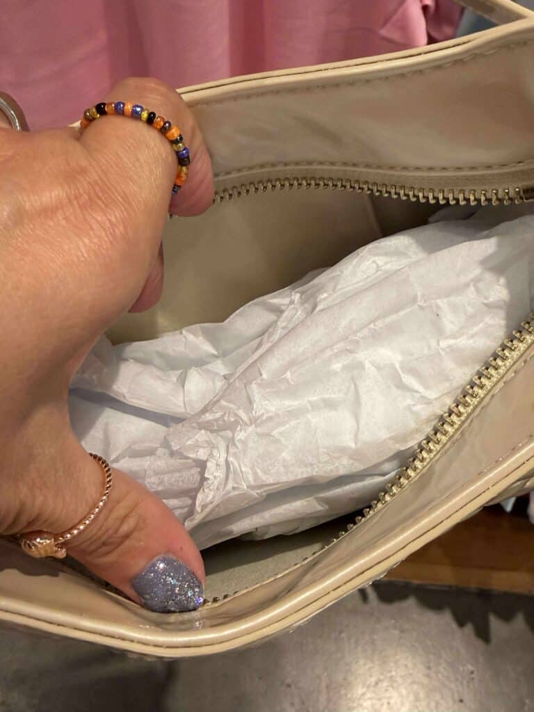 Beige handbag with tissue paper inside, zipper detail, and hand with colorful beaded ring.