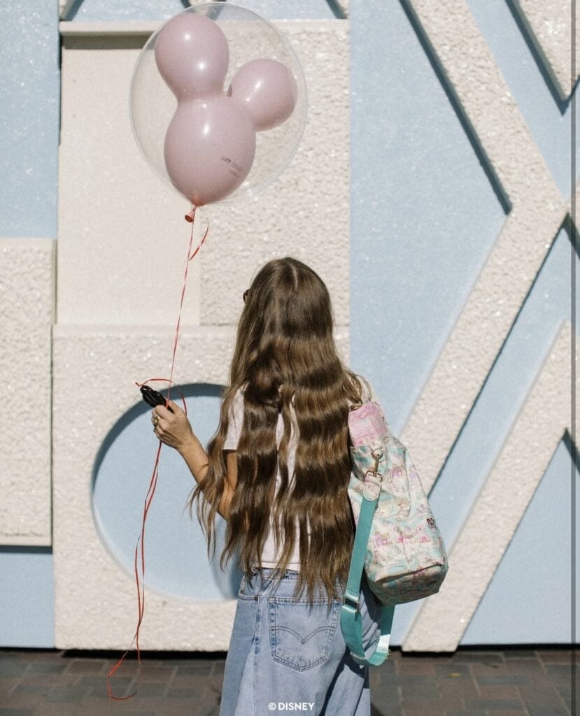 Pink Mickey Mouse balloon, girl with backpack, Disney fashion, casual style at Disney park, long wavy hair.