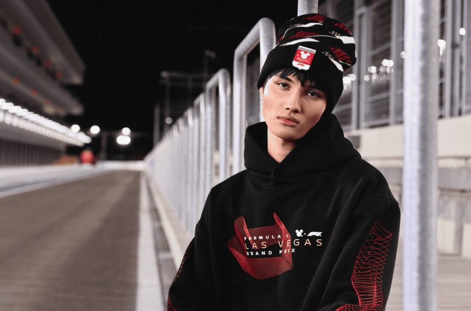 Stylish young man wearing Black Formula 1 Las Vegas hoodie and colorful fleece beanie at a night racetrack.