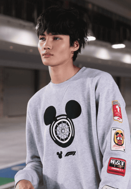 A young man wearing a gray Mickey Mouse graphic sweatshirt with motorsport patches, casual style, in an indoor setting.