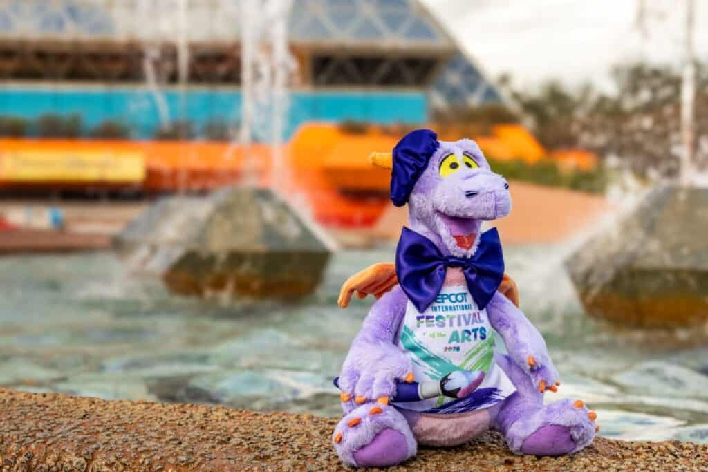 Colorful Disney dinosaur plush with festival-themed attire at an amusement park fountain, showcasing Disney merchandise and collectibles.