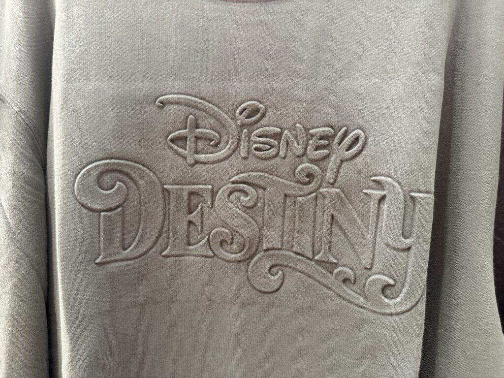 Close-up of embroidered Disney logo on beige fabric sweatshirt or hoodie.