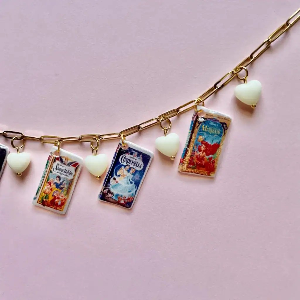 Close-up of a Disney-inspired charm necklace featuring miniature princess book charms and white heart pendants on a pink background.