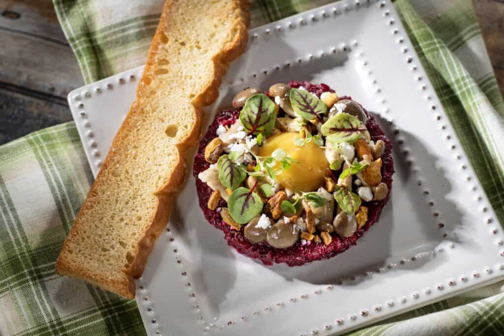 Vibrant beet tartare topped with microgreens, nuts, and a soft egg, served with toasted bread on a decorative plate.