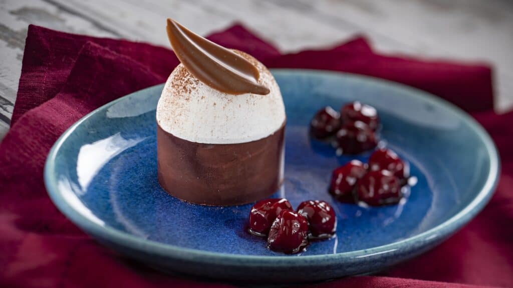 Rich chocolate mousse with garnished chocolate curl and fresh cherry topping on blue plate.