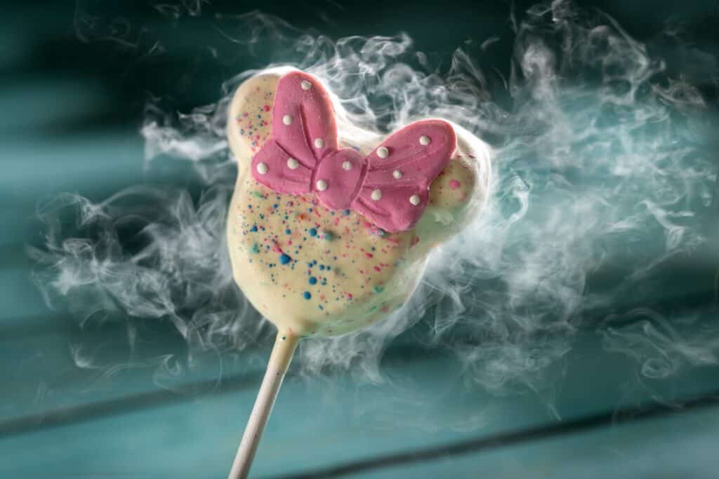 Colorful Mickey Mouse-shaped chocolate lollipop with pink butterfly decoration and vibrant sprinkles, surrounded by smoke effect.