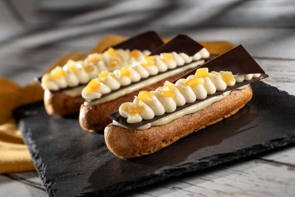 Delicious chocolate eclairs topped with white chocolate and mango chunks on a black slate plate. Perfect dessert idea.