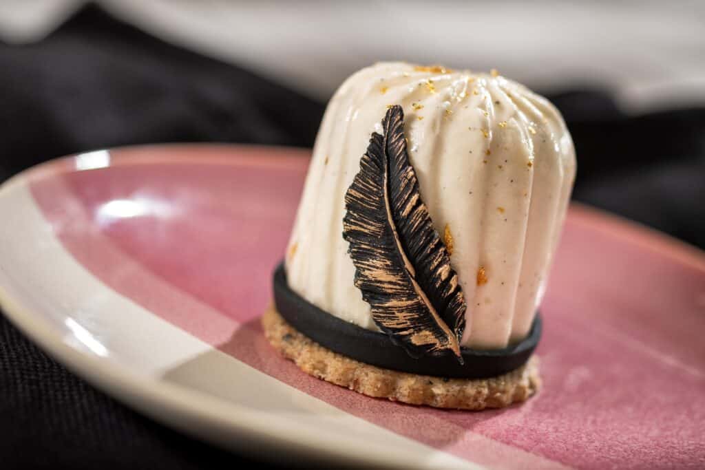Delicious Disney-inspired mousse dessert decorated with edible gold, chocolate feather, and served on a stylish pink plate.