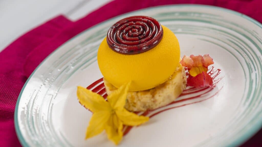 Colorful Disney-themed dessert with yellow mousse, red berry topping, and floral garnish on a stylish plate.