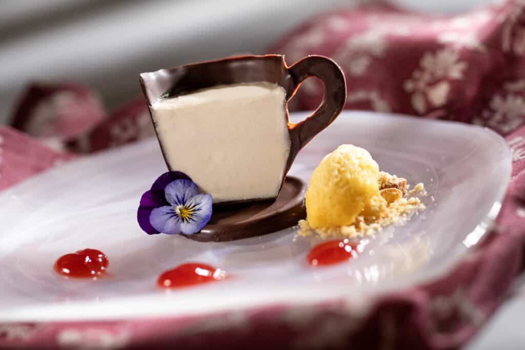 Delicious coffee mug cake with chocolate heart-shaped handle, edible flowers, and yellow sorbet on a decorative plate.