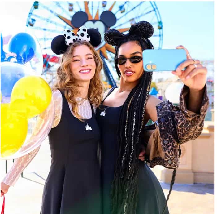 Two women wearing Minnie Mouse ears take a selfie at Disneyland park with a Ferris wheel in the background.