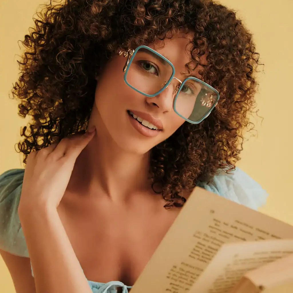 Stylish woman with curly hair and blue glasses reading a book, smiling happily.