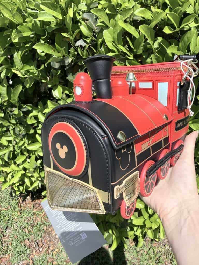 Red Disney train backpack with Mickey Mouse details, held outdoors against green foliage.