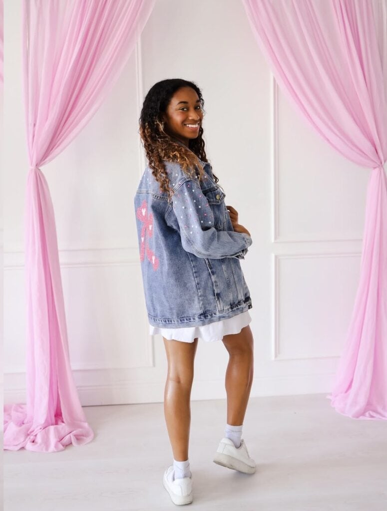 Girl wearing a denim jacket with pink heart patches, white dress, and sneakers in a pink curtain set.