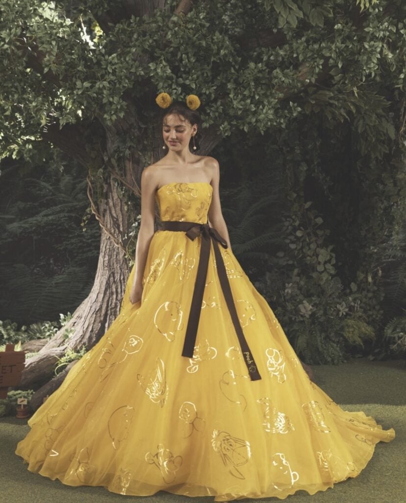 Woman in yellow princess gown with black ribbon, outdoor garden setting.