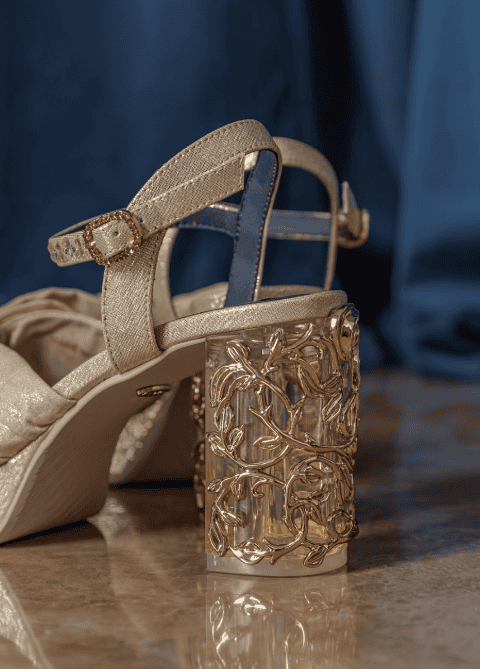 Stylish gold high-heeled sandal featuring a detailed transparent heel with floral accents.