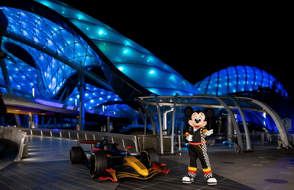Mickey Mouse standing next to a race car at a Disney-themed night event with modern architecture.