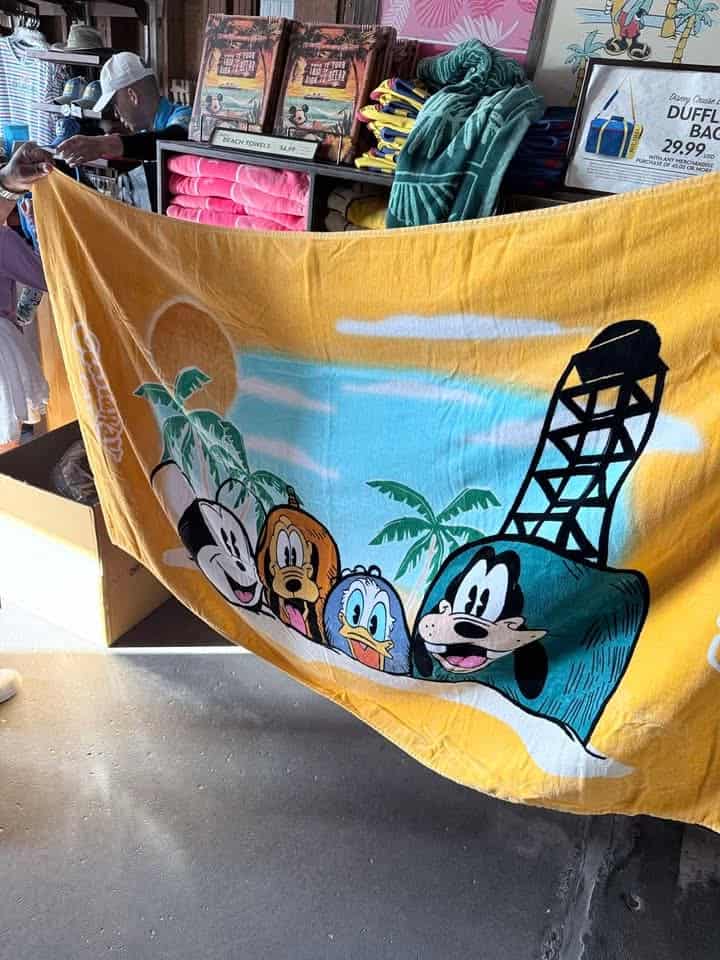 Beach towel featuring Mickey Mouse, Minnie Mouse, Donald Duck, and Goofy on a tropical beach scene.