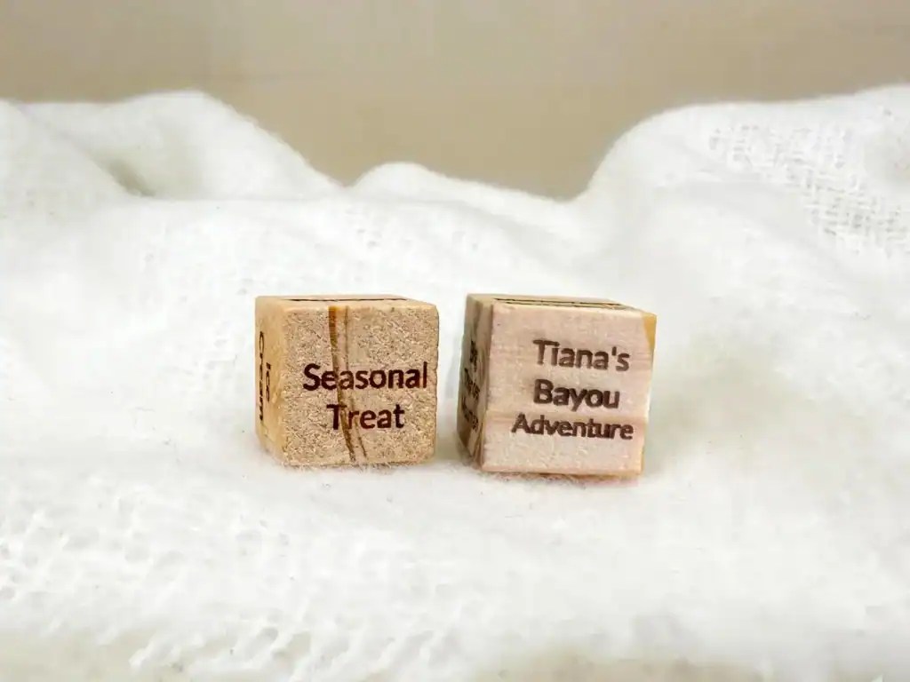 Wooden dice with "Seasonal Treat" and "Tiana's Bayou Adventure" text on white fabric.