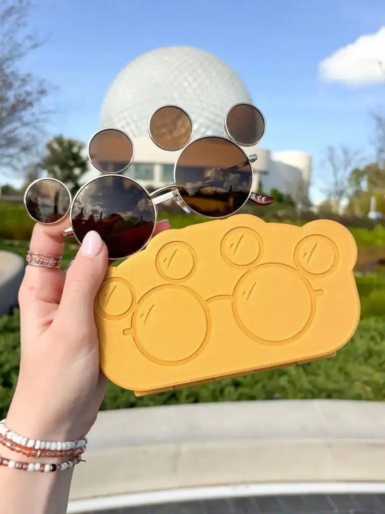 Mickey Mouse-shaped sunglasses held outdoors with Epcot ball in background.