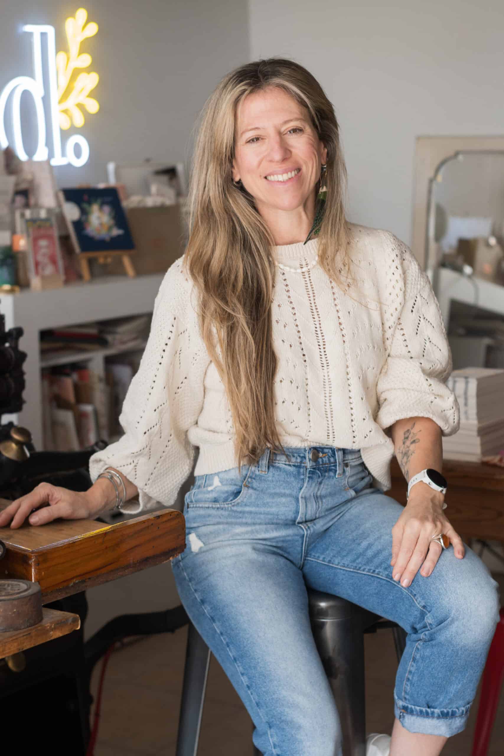 Artesana de papelería en letterpress en un taller creativo, sonriente, con ropa casual y cabello largo, rodeada de materiales de arte y diseño en su espacio de trabajo.