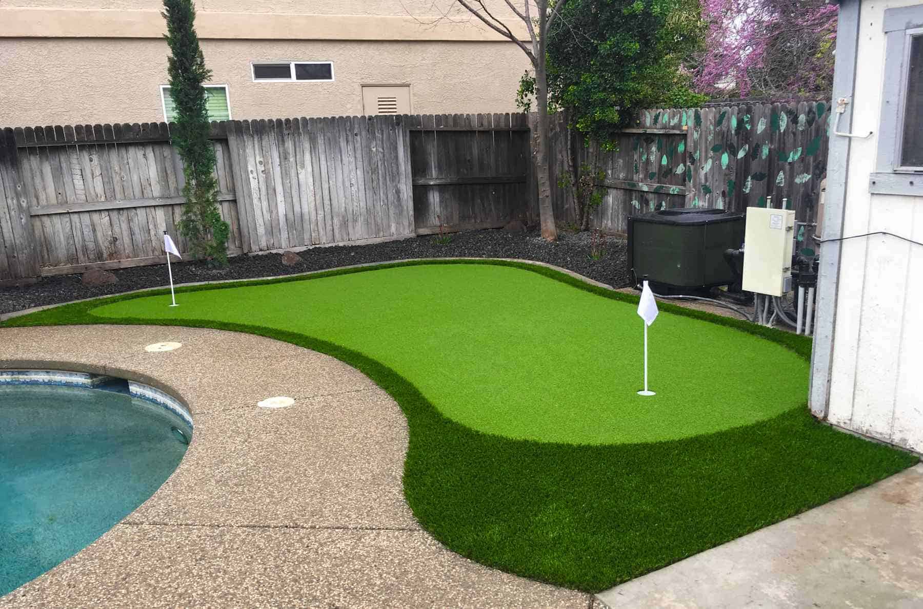 A vibrant artificial turf putting green with two flags sits beside an exposed aggregate pool deck in a Rio Vista backyard