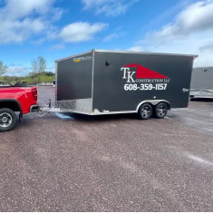 Black enclosed trailer with "TK Construction" logo, parked on gravel, showcasing quality trailer options at I39 Supply.