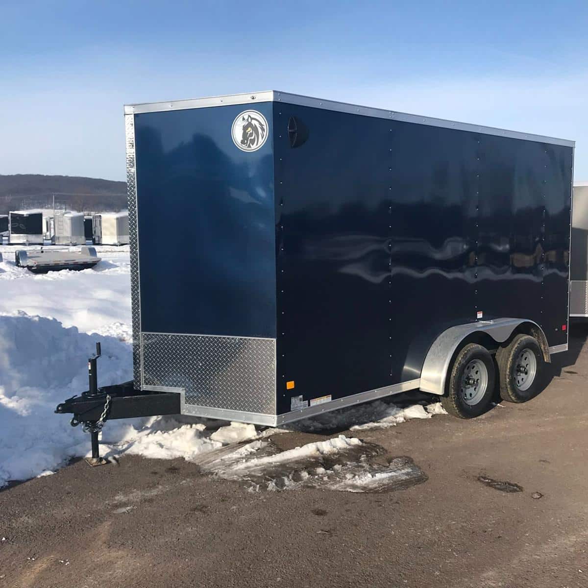 Durable navy blue enclosed cargo trailer with dual axles, ideal for transporting equipment and goods, parked on a paved lot with snow in the background.