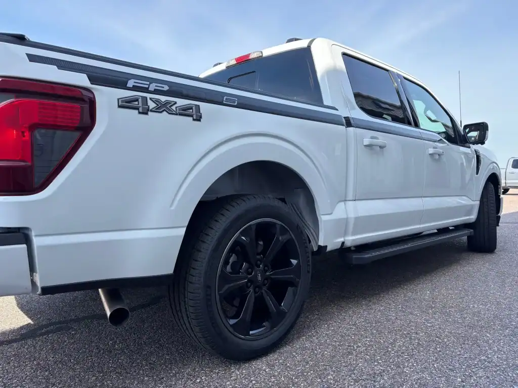 White Ford F-150 pickup truck with black wheels and 4x4 decal, parked on asphalt at I39 Supply trailer dealership.