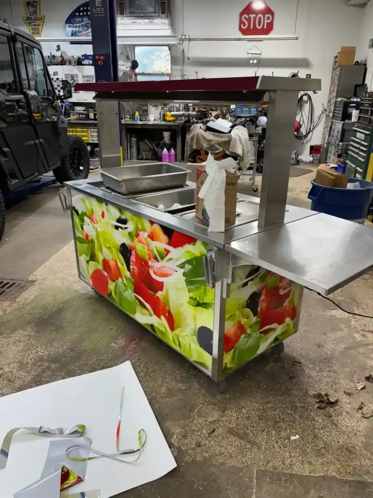 Commercial mobile food cart with colorful vegetable graphics, stainless steel surfaces, and overhead canopy, ideal for food vendors and catering businesses.