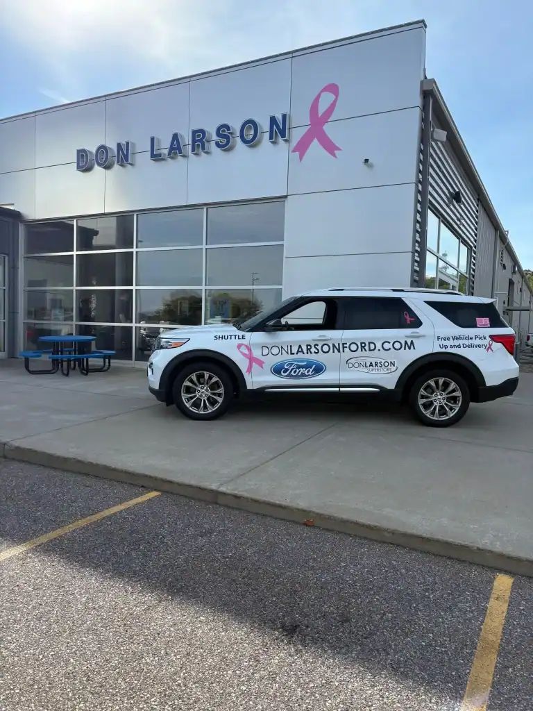 Exterior view of Don Larson Ford dealership with a promotional vehicle parked outside, showcasing the dealership's branding and pink ribbon awareness symbol.