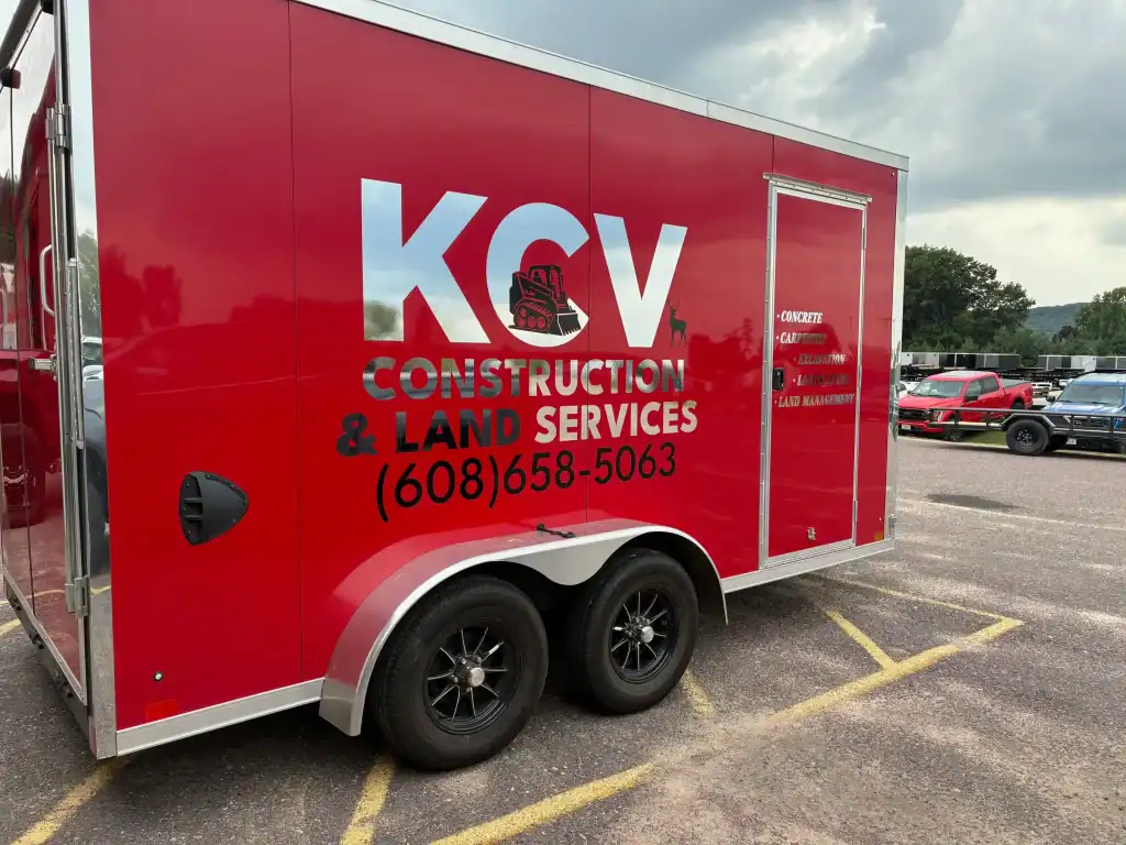 Construction & Land Services trailer with KCV logo, contact number, and branding, parked in a lot with other vehicles visible in the background.