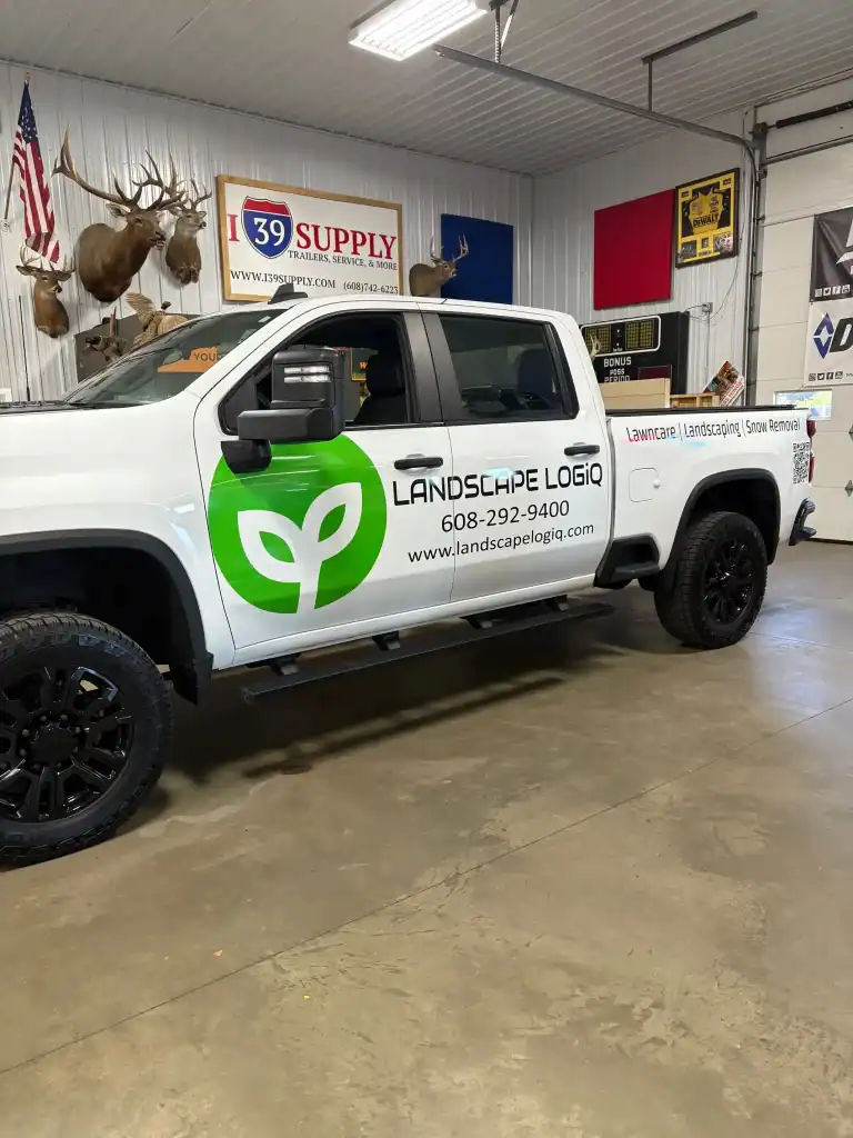 White pickup truck with Landscapes Logo and contact info parked inside a dealership with hunting trophies and signage in the background.