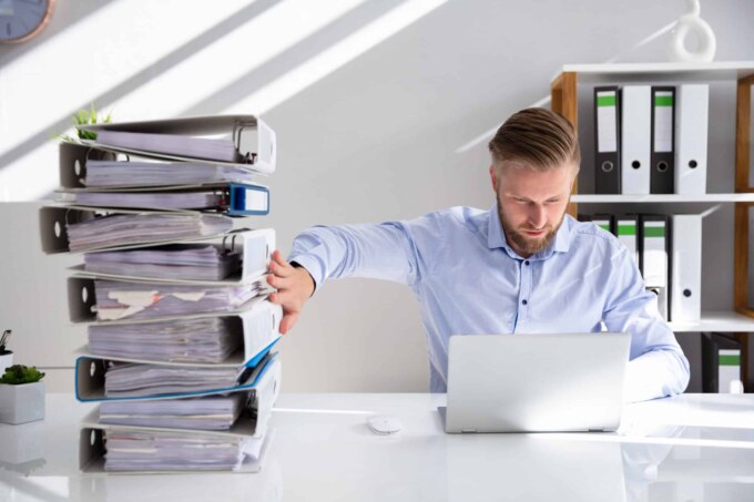 Man overwhelmed with stacked files and a laptop at a cluttered office desk.