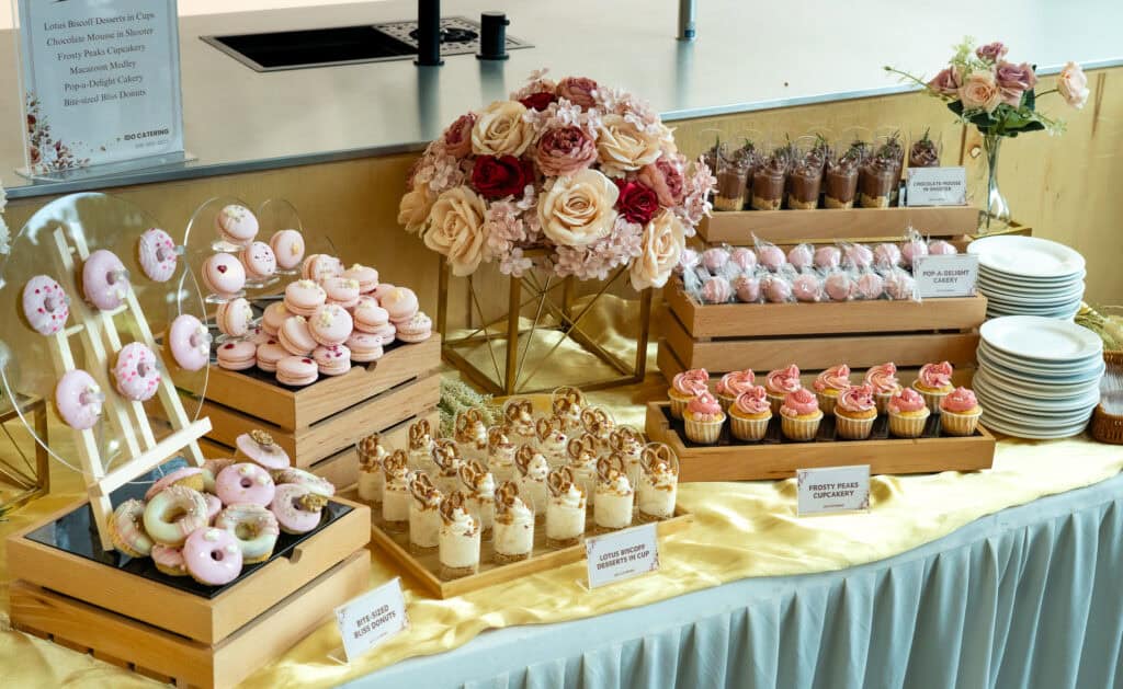 Wedding lunch catering featuring desserts in wooden crates and plates such as bite sized bliss donuts, lotus biscoff in cups, frosty peaks cupcakery, pop-a-delight cakery and pink macaroons by idocatering