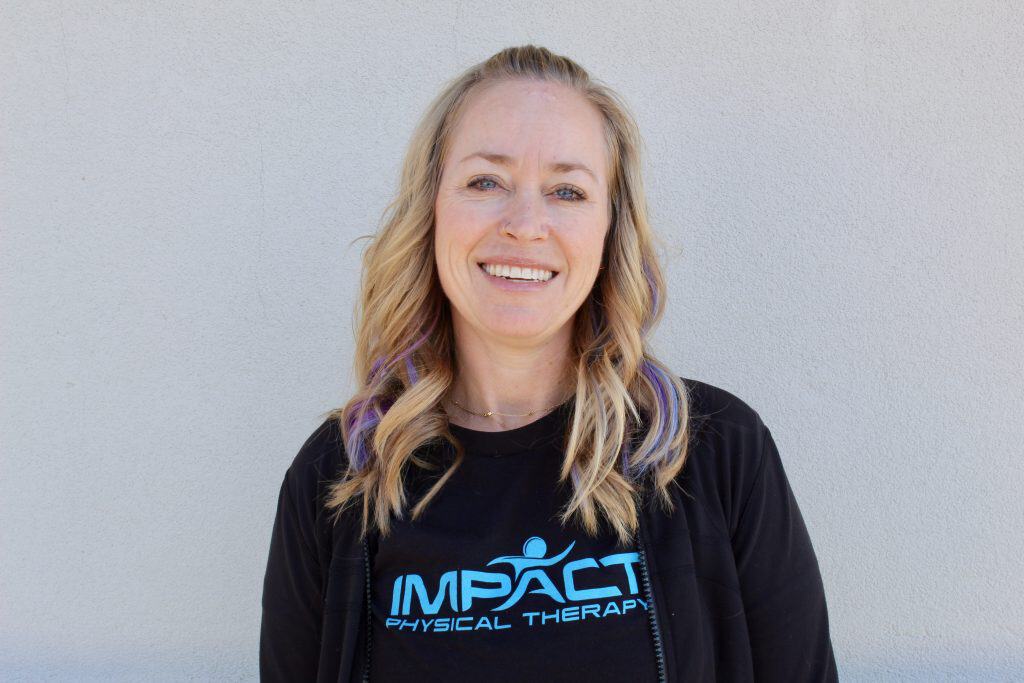 Smiling woman in Impact Physical Therapy shirt against neutral wall.