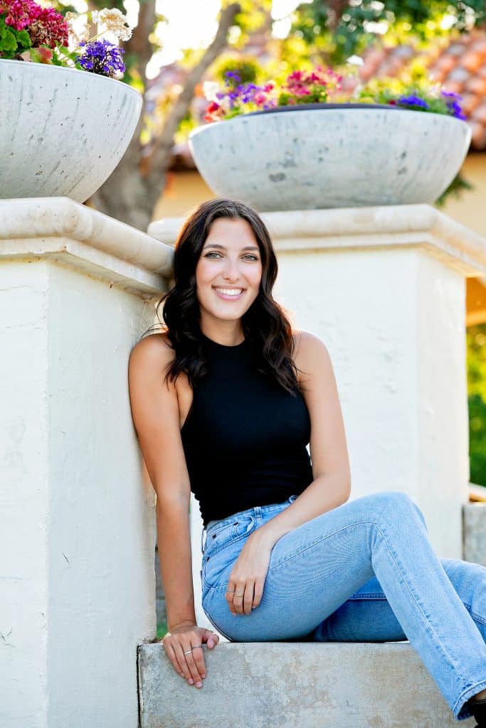 Smiling young woman sitting outdoors with flowers and trees in background.