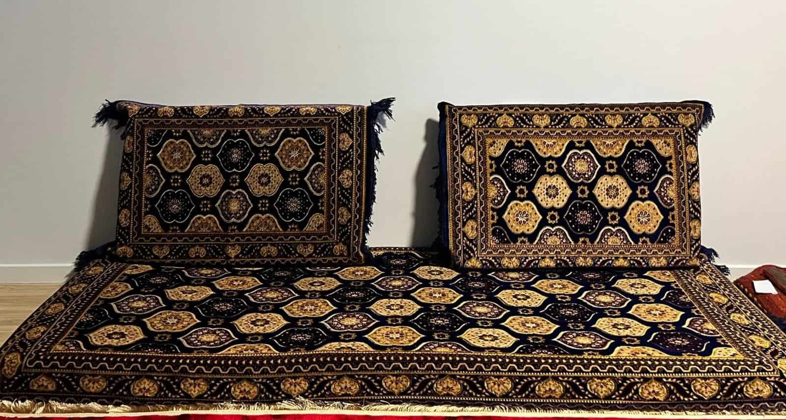 Two Cushioned Items With Black And Gold Geometric Patterns On A Matching Mat Against A White Wall.