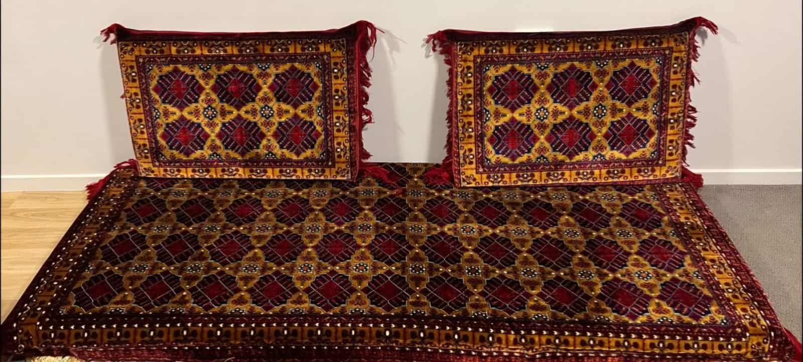 Two Ornate Cushions With Fringe On A Matching Geometric Carpet In Red, Yellow, And Blue Against A Wall.