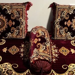 Plush cushions and bolster on an ornate rug with red, gold, and black floral patterns.