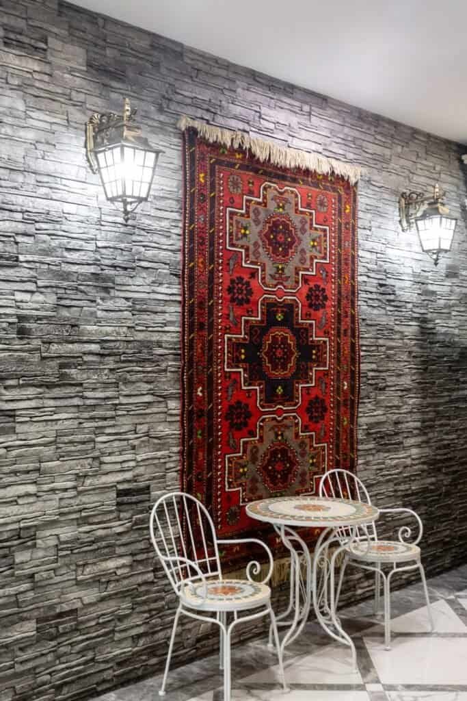 Afghani Handmade Rugs - The Fusion Of Heritage, Art, And Modern Decor 5 Decorative Scene With Red Tapestry On Stone Wall, Mosaic Table, Two Chairs, And Wall Lamps.