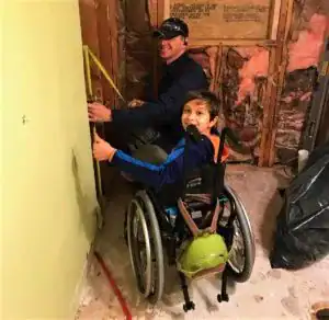 A child in a wheelchair gives a thumbs up while an adult measures a wall in a room under renovation with exposed insulation.